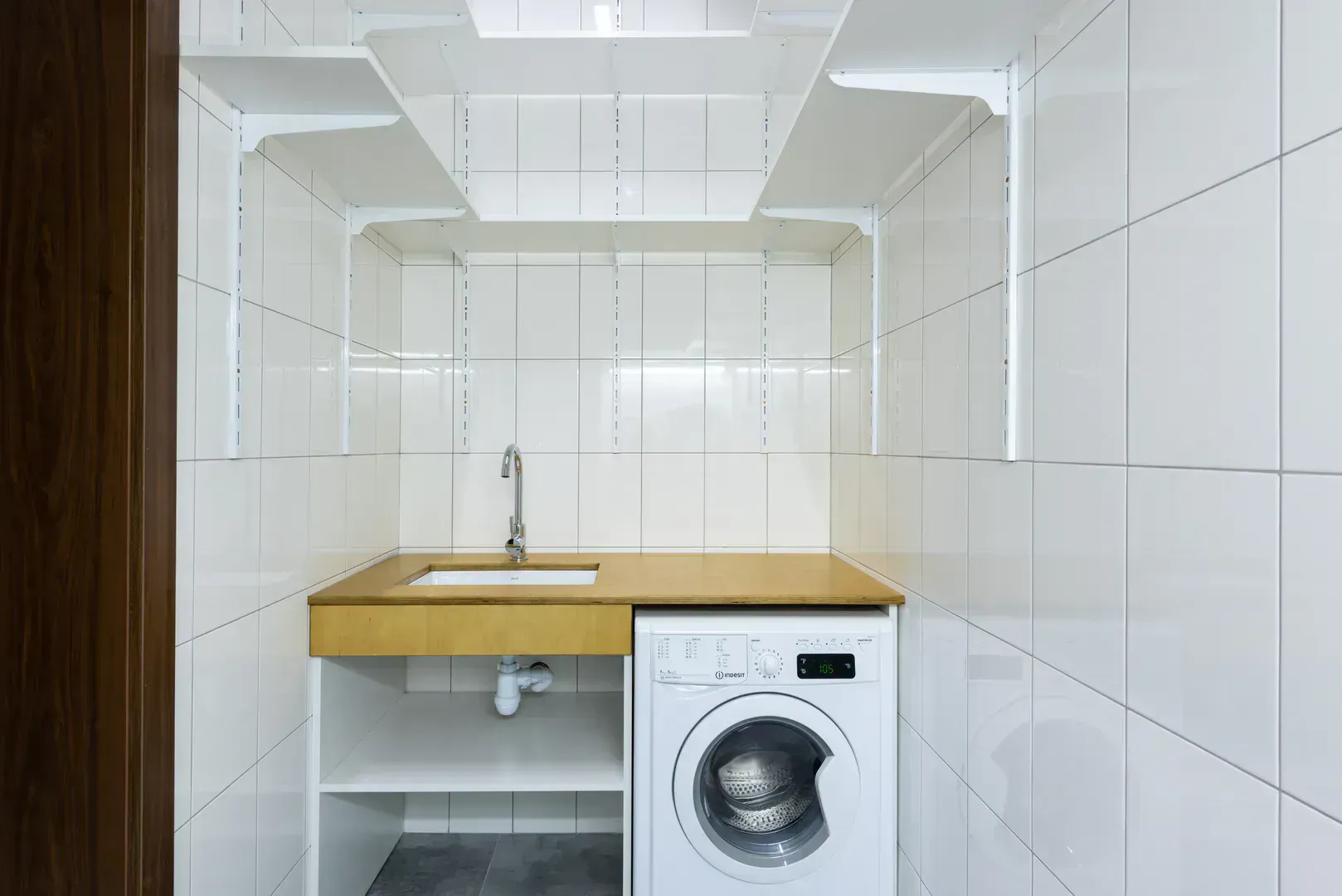 Washing machine in a white tiled laundry alcove with built-in shelves above the hookup