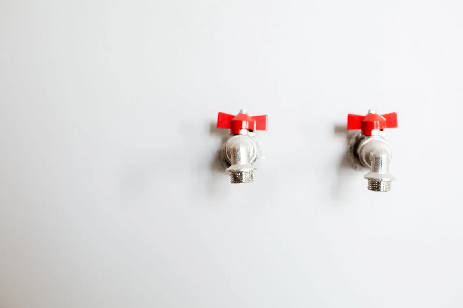 Two washing machine supply valves with red handles on a laundry room wall, where screw-on water hammer arrestors thread on